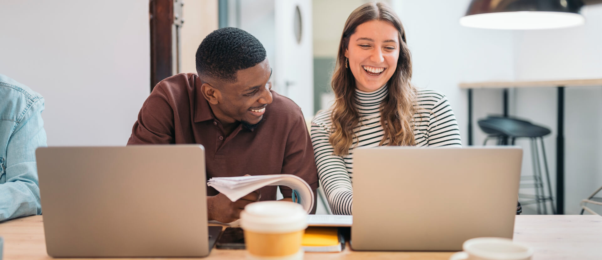 Two colleagues laughing together at a desk with laptops and a coffee cup