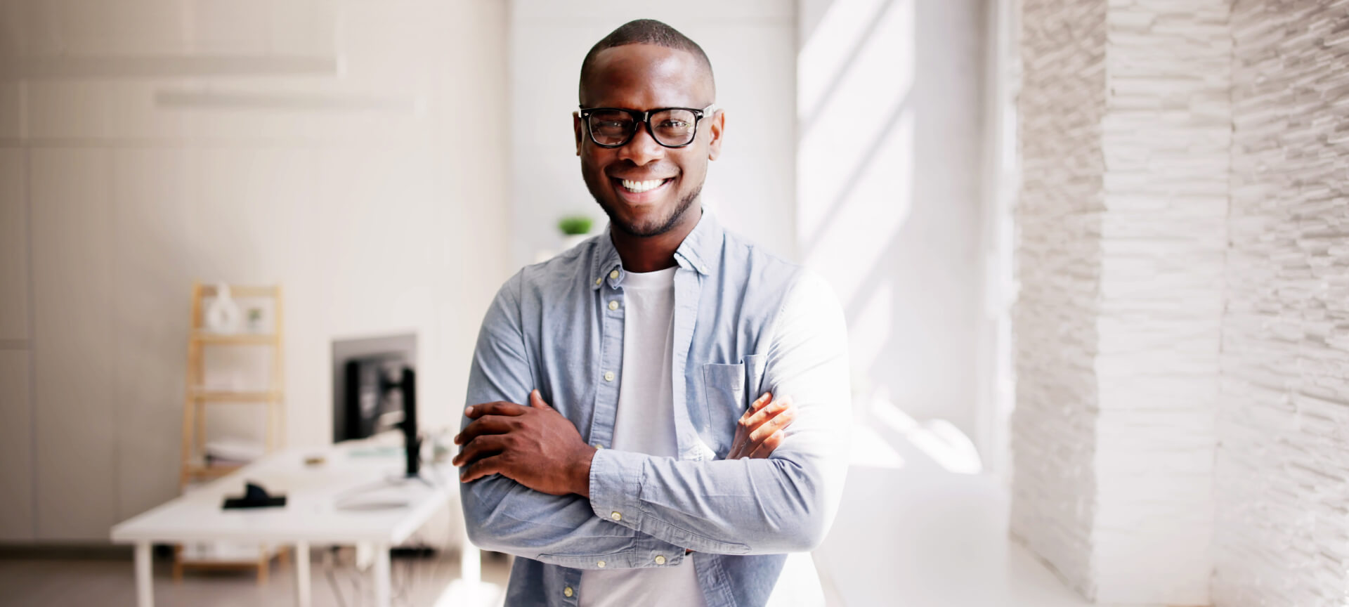 Smiling student with glasses and a denim shirt standing with arms crossed in an office