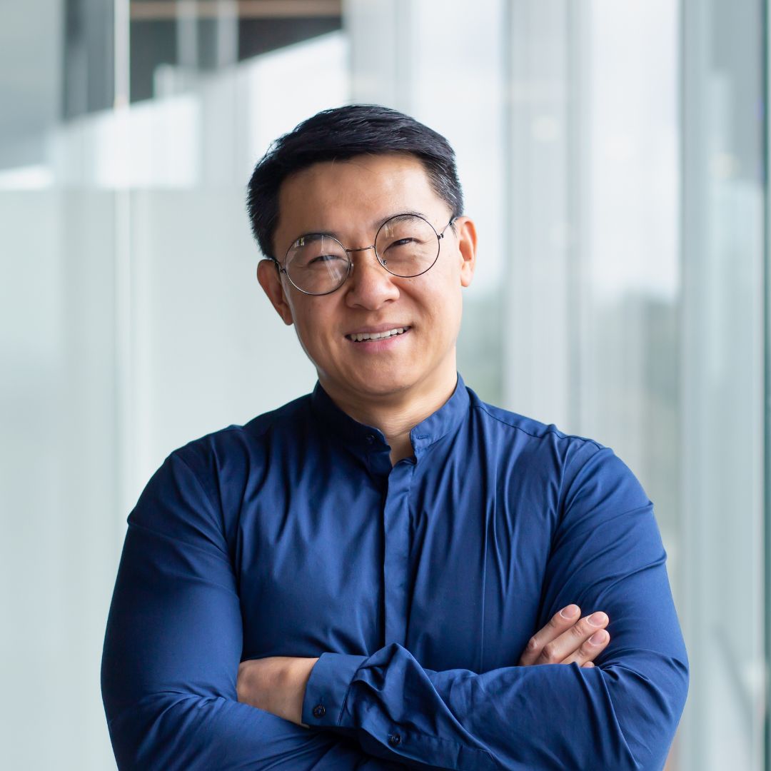 Smiling experienced man with round glasses and a blue shirt standing with arms crossed in an office