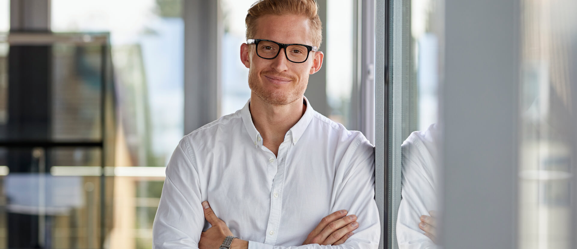 Smiling experienced professional with glasses and a white shirt leaning by a window, arms crossed