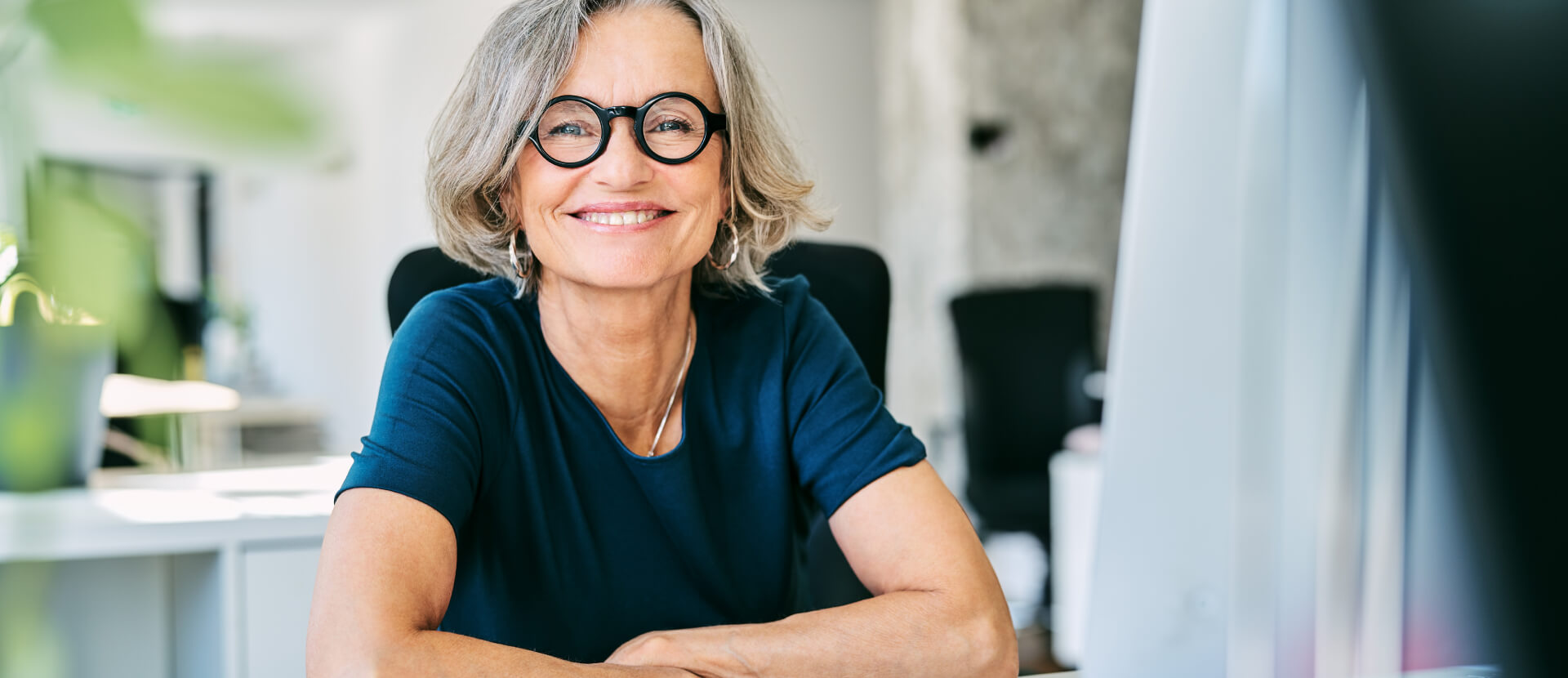 Smiling female manager with grey hair and round glasses sitting at her office desk