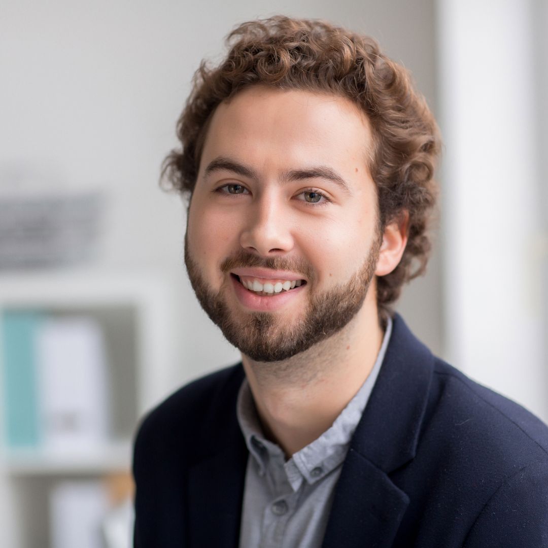 Smiling student with curly hair and a beard in a navy blazer in a bright office