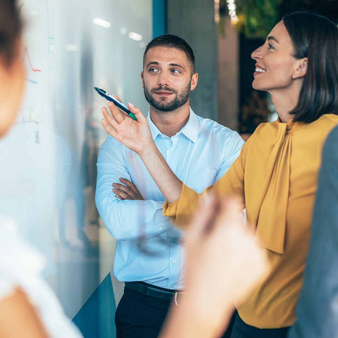 Smiling female colleague in a yellow blouse explaining at a whiteboard as a colleague listens