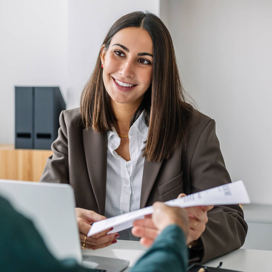 Smiling woman in a blazer in a meeting at a desk as a document is handed over