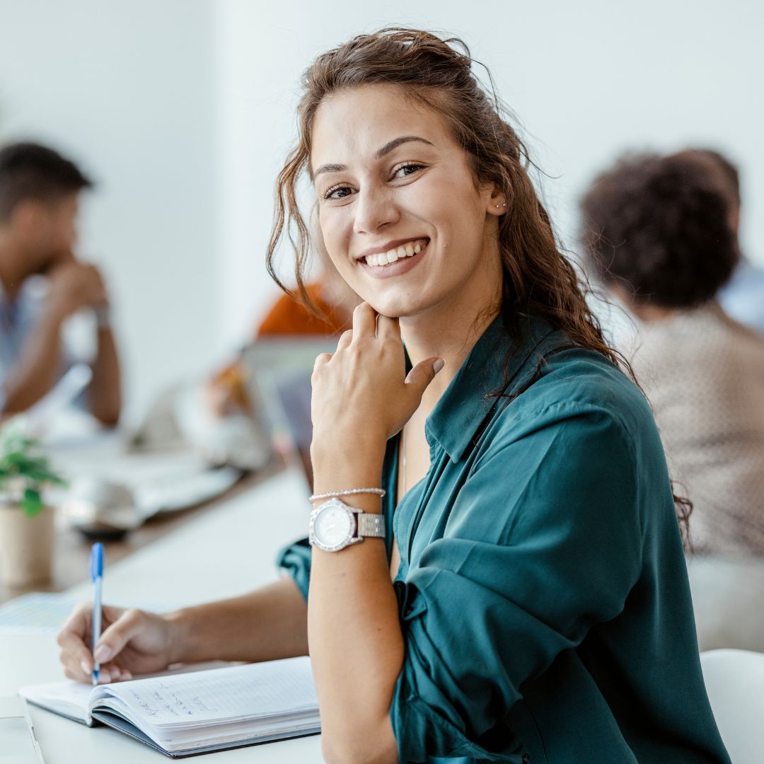 Smiling young woman in a green blouse taking notes at a desk in an office