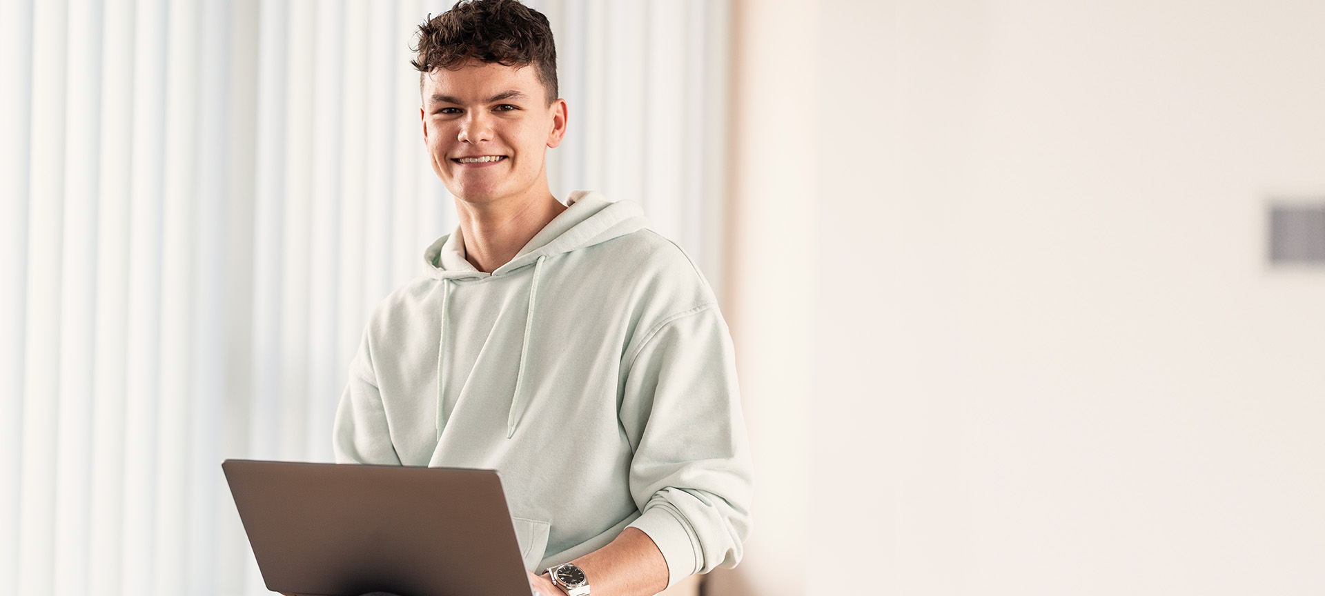 Lächelnder Auszubildender mit Locken im mintfarbenen Hoodie arbeitet am Laptop