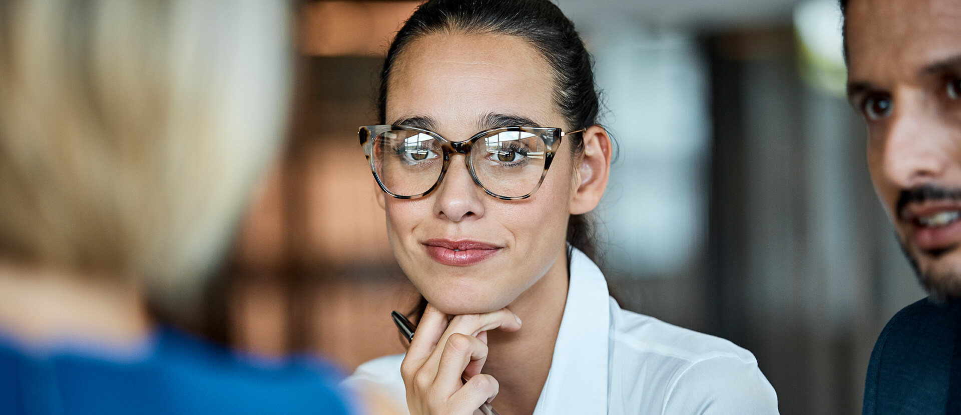 Lächelnder Kollege mit Brille und lächelnde Kollegin im Büro, dahinter arbeitet das Team