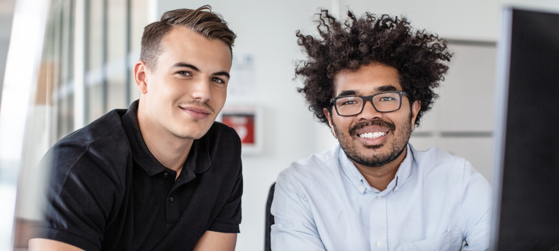 Zwei lächelnde Studenten, einer im schwarzen Poloshirt, einer mit Locken und Brille, im Büro
