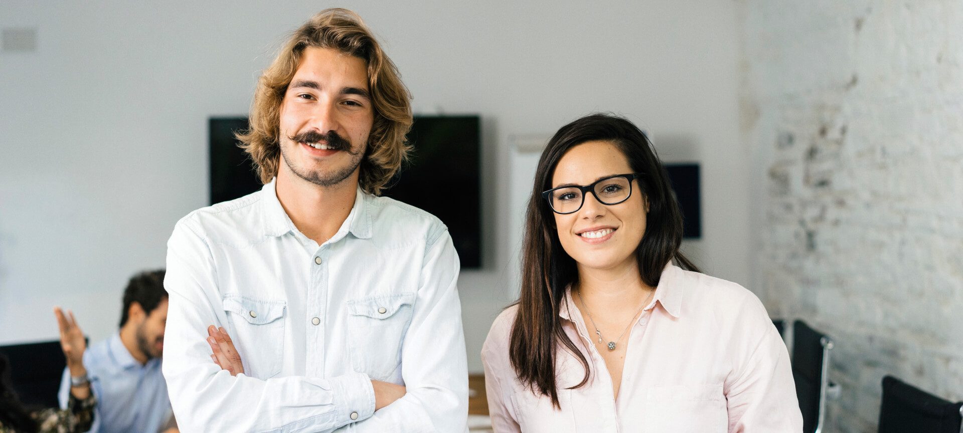 Zwei lächelnde Berufseinsteigende, er mit Schnurrbart, sie mit Brille, stehen im Büro